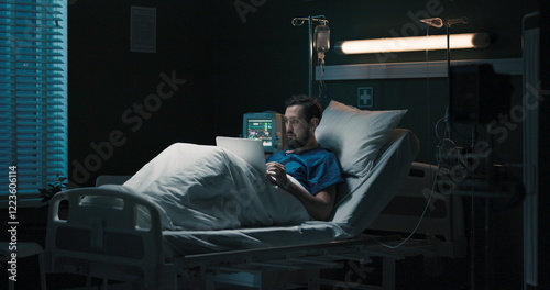 Concentrated young beard patient in hospital bed with laptop works online.