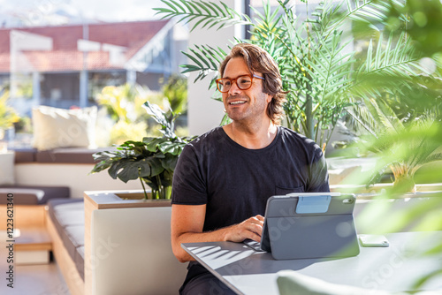 A smiling happy man is sitting at a table with a tablet in front of him working