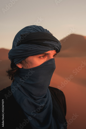 Caucasian male tourist wearing blue turban and shemagh exploring erg chebbi dunes in merzouga, sahara desert, morocco, during golden hour