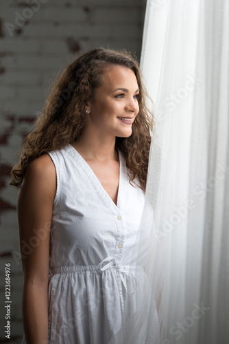 Sexy young poses in studio wearing white dress stands at window photo