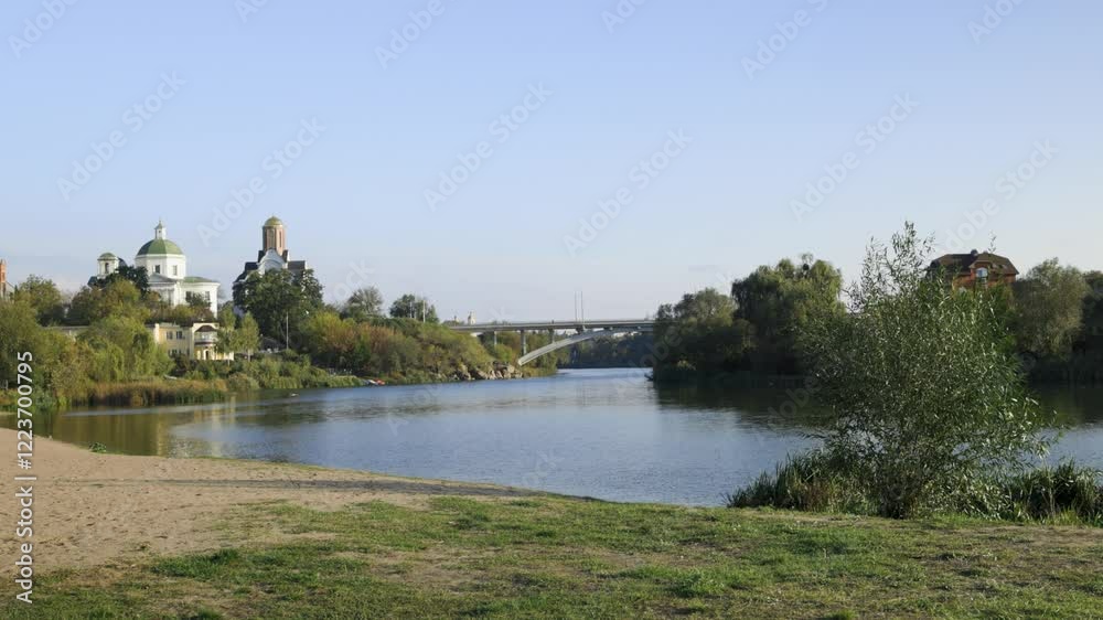 A peaceful riverside scene with lush greenery, a bridge, and historic buildings, including a church with a dome. The calm water reflects the serene atmosphere of the sunny day. A peaceful riverside scene with lush greenery, a bridge, and historic buildings, including a church with a dome. The calm water reflects the serene atmosphere of the sunny day.