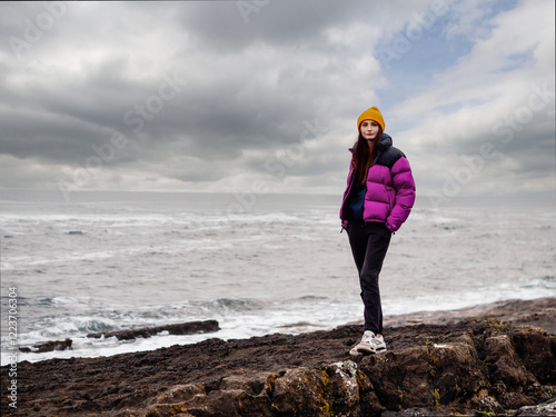 A woman stands on a stone coast overlooking the ocean. She is wearing a pink jacket and a scarf. The scene is peaceful and serene, with the ocean as a backdrop. Travel in Ireland in winter photo