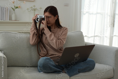 Young Asian woman sitting cross-legged on sofa in living room holding camera working selling stock photos living room at home startup and working from home concept