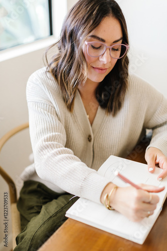 Woman in glasses writing in a notebook at a table near a window.