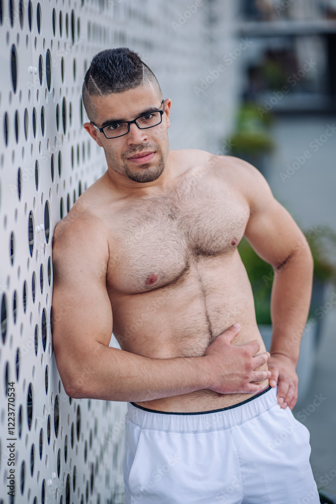 Shirtless sportsman with glasses and a striking mohawk hairstyle posing confidently against a modern perforated metal wall in russia