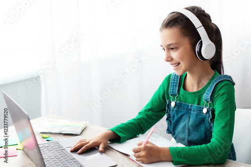 Studying At Home. Cheerful schoolgirl doing homework and typing on keyboard, sitting at table using laptop, looking at screen, writing essay or making test, taking notes in textbook, free copy space photo