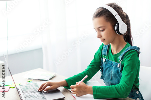 Educational Lecture Concept. Portrait of diligent teen girl in wireless headphones sitting at table using laptop, writing in copybook taking notes in notebook, listening to teacher, free copy space