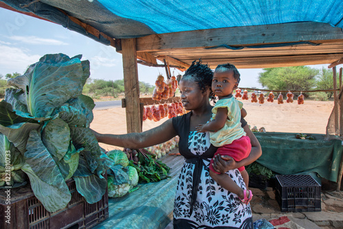 African woman vendor holding baby selling vegetables from a wooden shack stall in the city, on the side of the road, stall fresh produce onions, tomatoes, cabbage, representing rural commerce