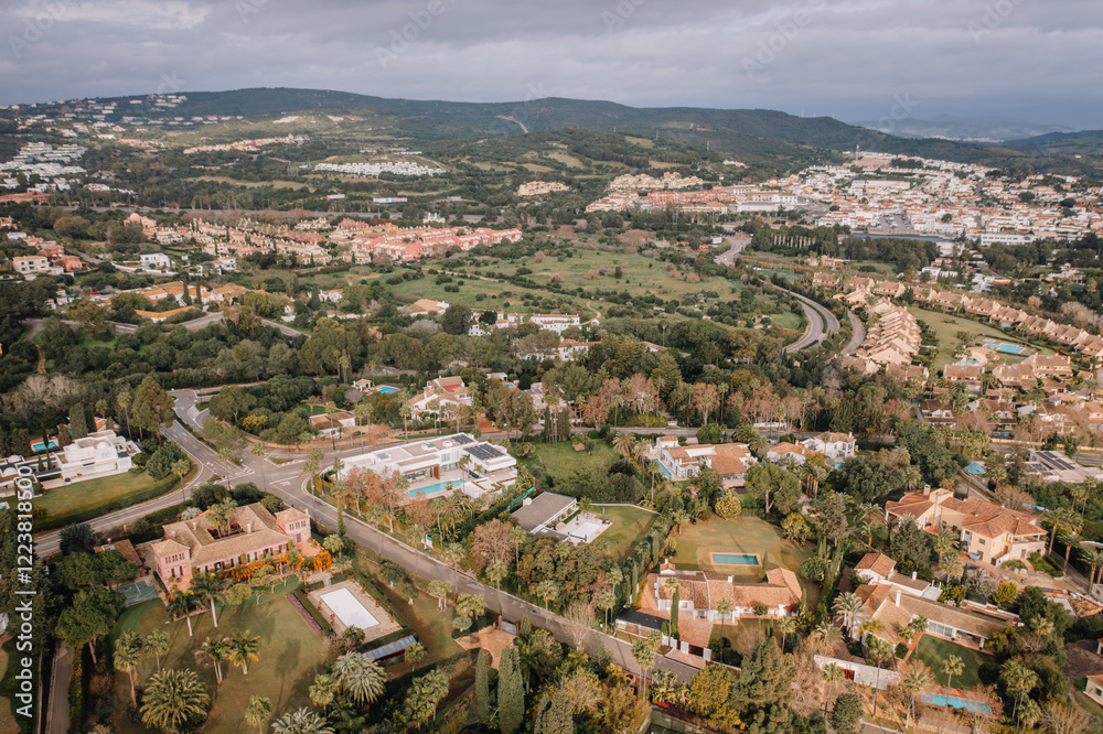 Obraz premium Aerial view of a luxury residential area with villas, private pools, gardens, and nearby greenery surrounded by scenic hills and urban landscapes.