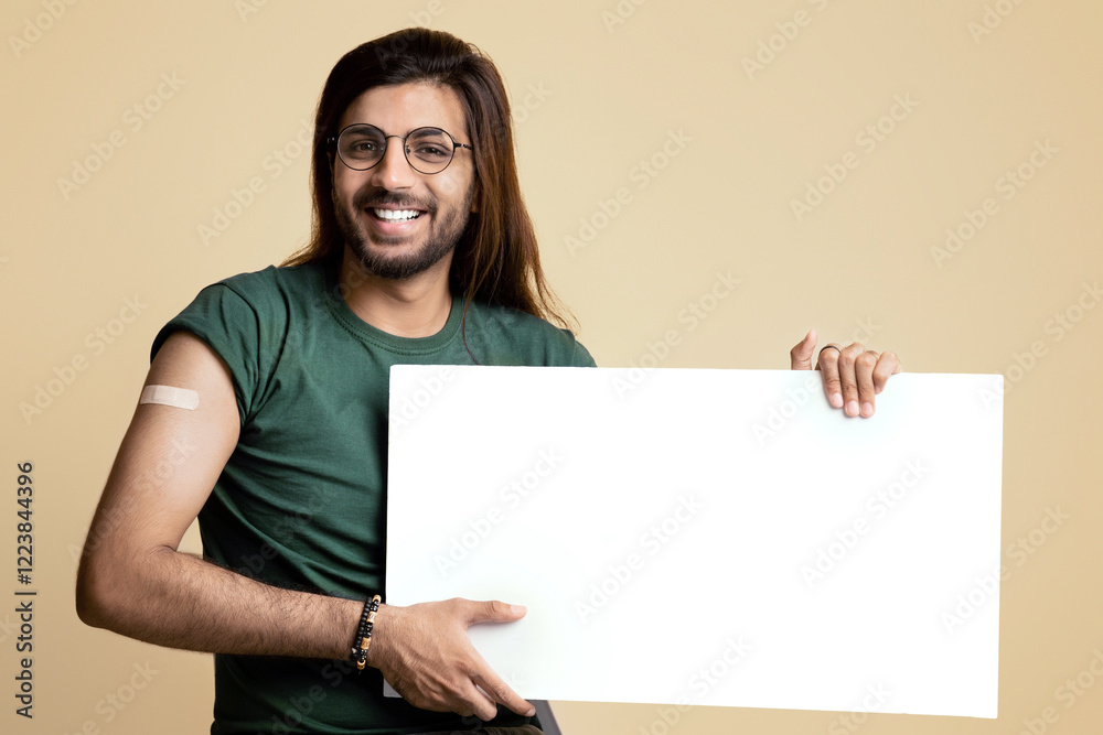 Healthy arab guy got vaccinated against coronavirus, sitting on chair over beige studio background, showing adhesive plaster on his shoulder and empty horizontal board for text or advertisement Healthy arab guy got vaccinated against coronavirus, sitting on chair over beige studio background, showing adhesive plaster on his shoulder and empty horizontal board for text or advertisement