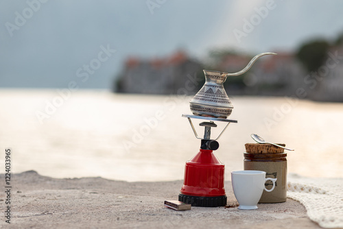 A traditional coffee pot sits on a small gas stove near the waters edge, with a cup and jar nearby. The calm surface of the water reflects the light as the sun rises, creating a peaceful atmosphere. photo