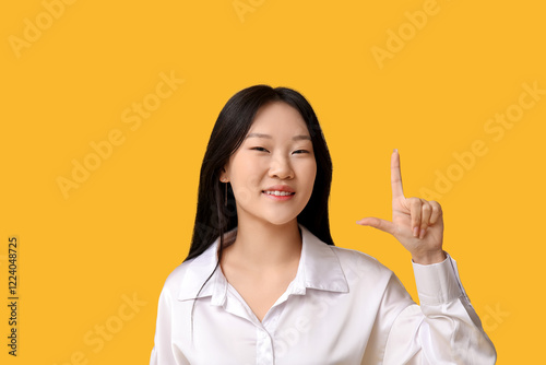 Beautiful young Asian woman pointing at something on yellow background