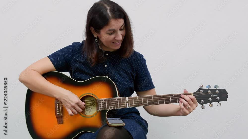 Guitarist practicing new chords on an acoustic guitar while using a smartphone app, set against a clean white wall in an indoor space, focusing on skill development and musical creativity Guitarist practicing new chords on an acoustic guitar while using a smartphone app, set against a clean white wall in an indoor space, focusing on skill development and musical creativity