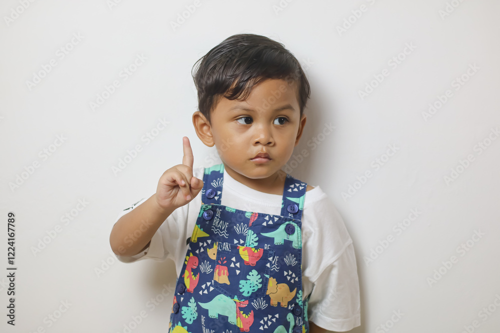 Cute smiling boy in plain white t shirt looking and pointing hand up in isolated studio light blue color background