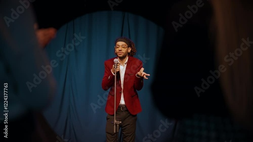 Stand-up show in trendy bar or club, visitors applauding to man on stage. Communication of speaker and audience, people enjoying speech of charismatic african american guy in red jacket, portrait photo