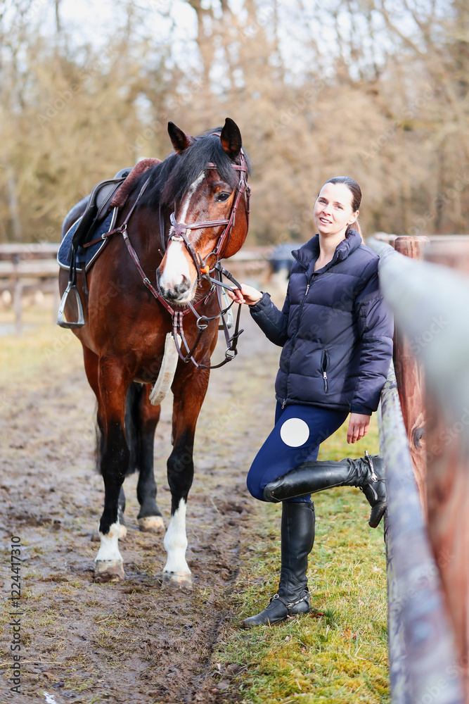 A young woman stands with her saddled horse model between the wooden fences of the outdoor paddocks in winter nature. A young woman stands with her saddled horse model between the wooden fences of the outdoor paddocks in winter nature.