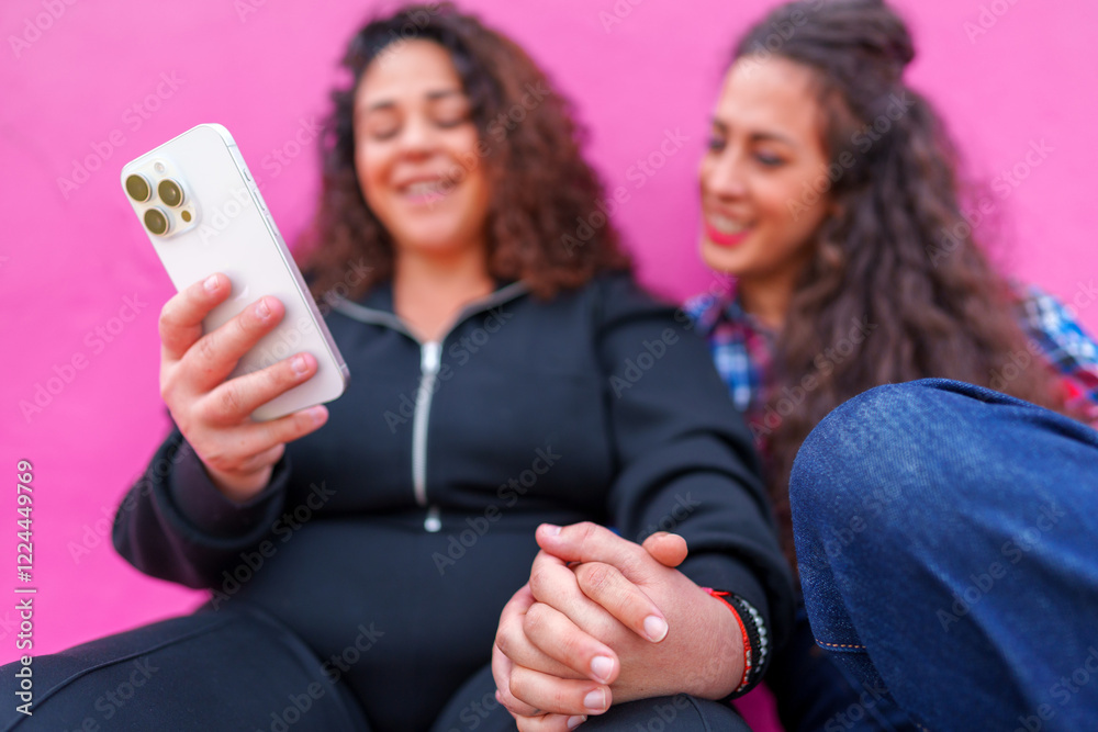 Pregnant woman connecting with friend, laughing while viewing smartphone content, seated against bright pink setting Pregnant woman connecting with friend, laughing while viewing smartphone content, seated against bright pink setting