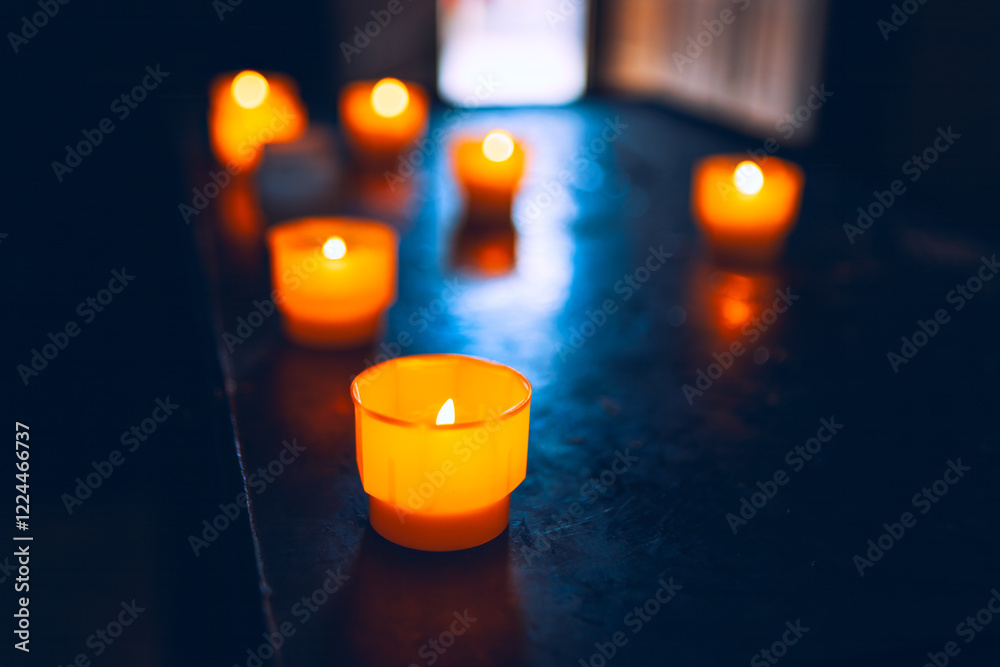 Dark surface with glowing orange candles. The candles emit a warm light, creating a cozy atmosphere. Light from a window can be seen in the background. Praying indoor the church