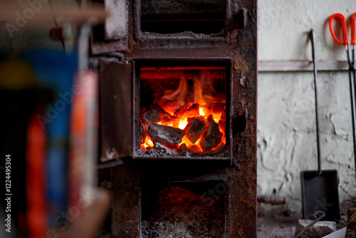 A rustic stove with a burning fire, a coal and firewood shovel and tongs standing nearby. Heating in a private house, fireplace, fire safety.