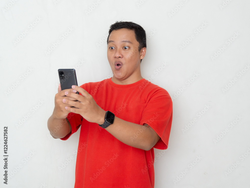Young Asian man in red shirt smiling and looking at his smartphone. Man shows joyful and happy expression while pointing to his mobile phone Young Asian man in red shirt smiling and looking at his smartphone. Man shows joyful and happy expression while pointing to his mobile phone