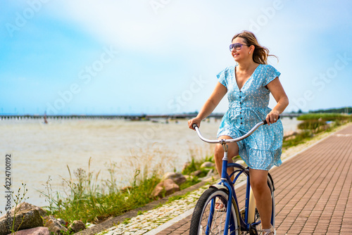 Active summer holidays. Mature woman wearing dress riding blue cruiser bike on bicycle path on seafront on sunny day. Front view