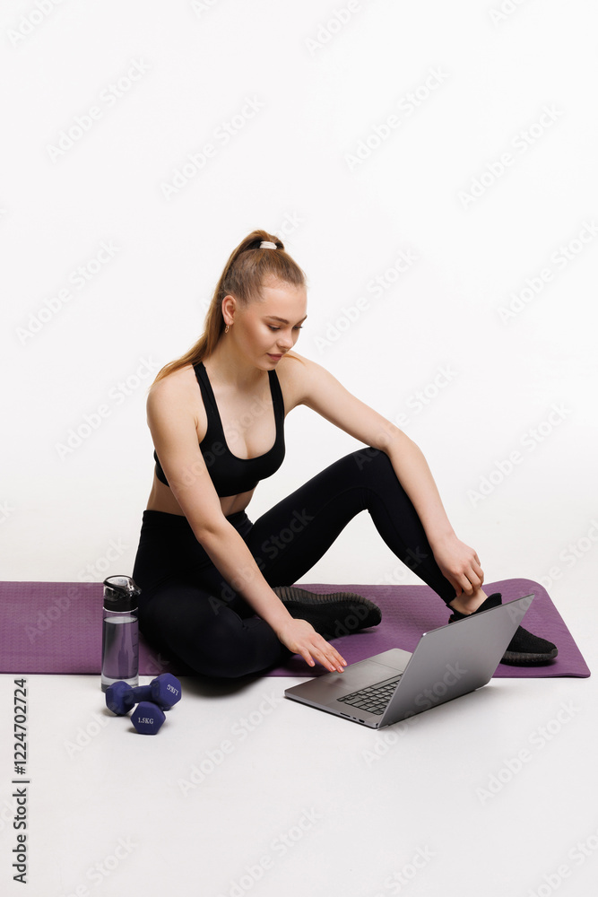 Fit woman in active clothes using laptop while sitting near bottle and fitness mat on white background Fit woman in active clothes using laptop while sitting near bottle and fitness mat on white background