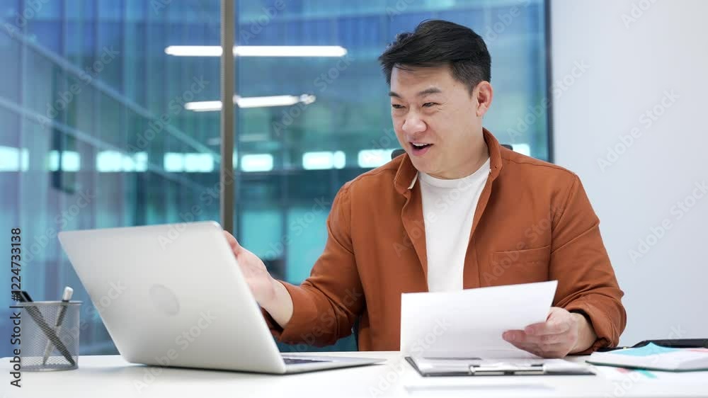 Happy asian businessman satisfied with financial results sitting at workplace in business office. Smiling financier looks at documents and laptop, celebrates success, rejoices at positive indicators