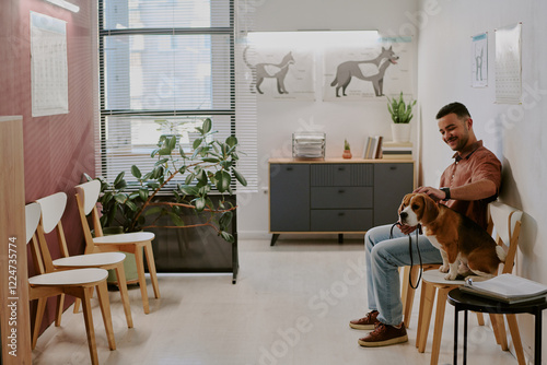 Man sitting in veterinarian's waiting room with dog on lap, surrounded by chairs and decor. Various animal posters adorning walls and plants adding a refreshing touch photo