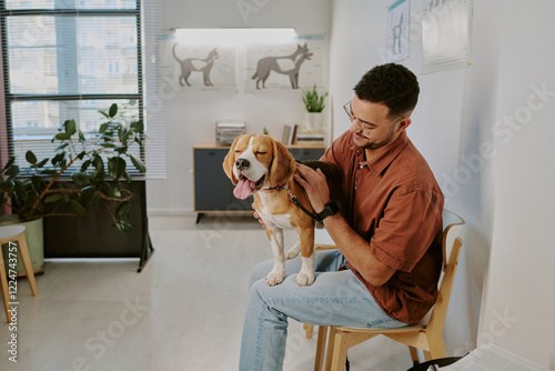 Smiling man sitting in veterinary clinic with his dog on his lap while they wait for consultation. Clinic setting with pet-related posters on wall and potted plants
