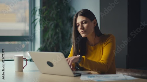 Young woman smiling while working on a laptop in a modern office, wearing a yellow sweater, radiating happiness and focus. Concept of positivity, productivity, and professional lifestyle