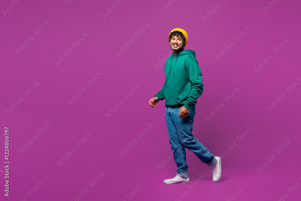 Full body side profile view young smiling happy fun Indian man he wear green hoody yellow hat casual clothes walk go look camera isolated on plain purple background studio portrait. Lifestyle concept. Full body side profile view young smiling happy fun Indian man he wear green hoody yellow hat casual clothes walk go look camera isolated on plain purple background studio portrait. Lifestyle concept.