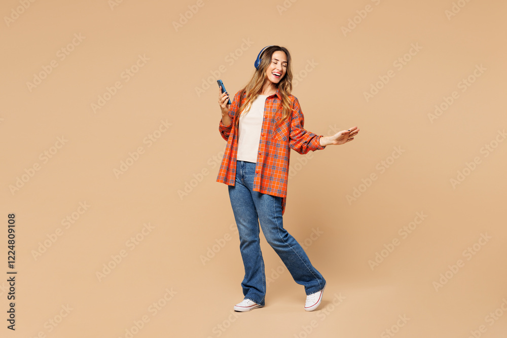 Fototapeta premium Full body young fun woman she wear orange shirt casual clothes listen to music in headphones use mobile cell phone isolated on plain pastel light beige background studio portrait. Lifestyle concept.
