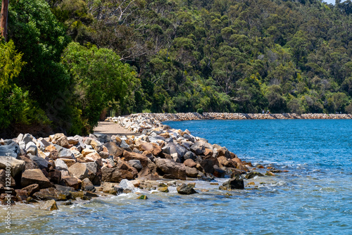 A rocky breakwater stretches along the pristine blue coastline, with large granite boulders protecting the shoreline from erosion. Australian coastal landscape at Kalimna Jetty Walk, Lakes Entrance. photo