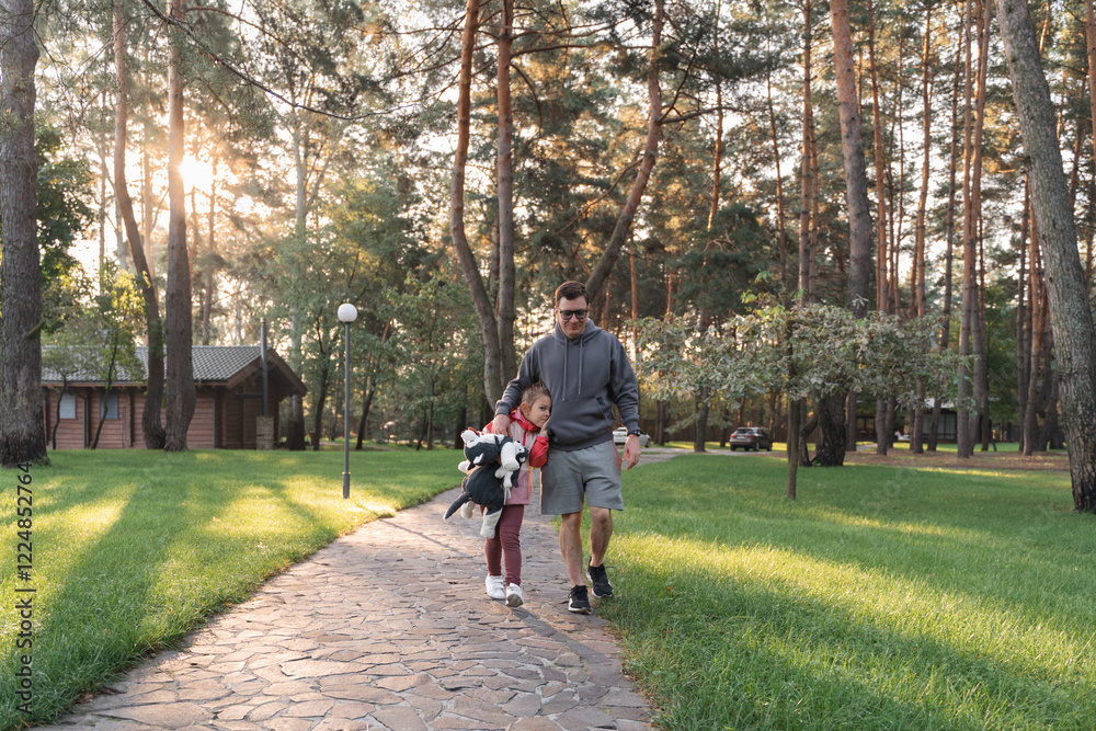 A young girl smiles brightly, holding a stuffed animal, while walking with her father in a green park A young girl smiles brightly, holding a stuffed animal, while walking with her father in a green park