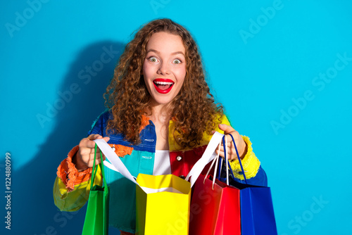 Excited young woman holding colorful shopping bags against vibrant blue background, demonstrating joy and surprises of retail therapy photo