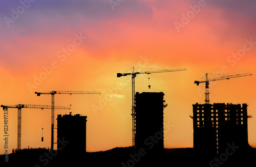 Fototapeta Naklejka Na Ścianę i Meble -  Silhouettes of tower cranes constructing a new residential building at a construction site against sunset background.
