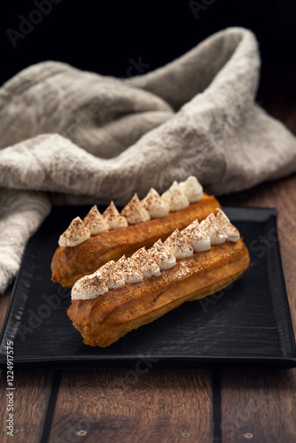 Two eclairs topped with whipped cream and dusted with cocoa powder are placed on a black rectangular plate. A blurred background includes a brown ceramic cup and a textured fabric