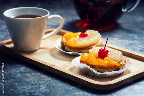A cup of tea and two pineapple upside-down cakes with cherries on top are placed on a wooden tray, with a teapot in the background