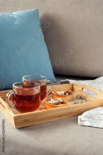 Two cups of tea on a saucer with two small tarts topped with white sprinkles, placed on a beige couch with blue pillows in the background