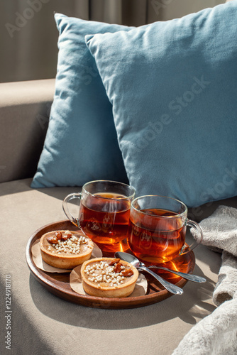 Two cups of tea on a saucer with two small tarts topped with white sprinkles, placed on a beige couch with blue pillows in the background