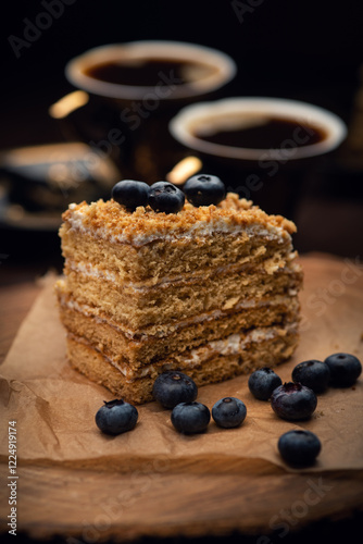 A slice of layered cake topped with blueberries sits on parchment paper, with two cups of coffee in the background, creating a cozy and inviting scene.