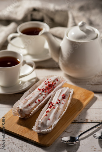 Two éclairs with white icing and red sprinkles on a wooden board, accompanied by a cup of tea and two spoons on a rustic wooden table.