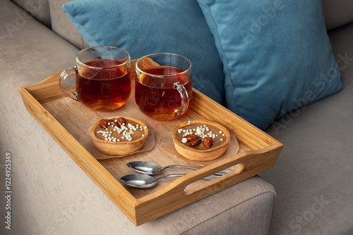 Two cups of tea on a saucer with two small tarts topped with white sprinkles, placed on a beige couch with blue pillows in the background