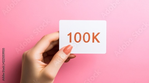 Woman s hand holding a white card with 100K in rust-orange numerals on a pastel pink background photo