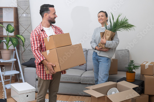 Settling In Abroad: A content and delighted couple, after relocating internationally, stands in their new living room with a pile of cardboard relocation service boxes, feeling at home.