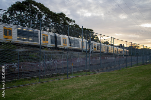 A stunning shot capturing a train beside the International Peace Park in Western Sydney, Australia. The illuminated cityscape meets a vibrant sky, with long trees in the backdrop.