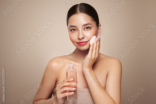 Smiling woman holds bottle with cosmetic tonic. Photo of attractive woman with perfect makeup on beige background. Beauty and skin care concept. Removing makeup