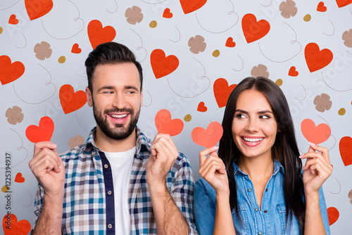 Love story of sweet, cheerful, positive, smiling couple in shirts showing small red paper hearts in two hands, looking to each other with eyes over grey background photo