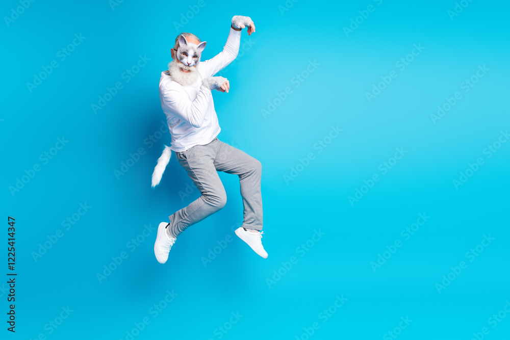 Obraz premium Elderly man in a fun outfit with cat mask jumping joyfully against a blue background