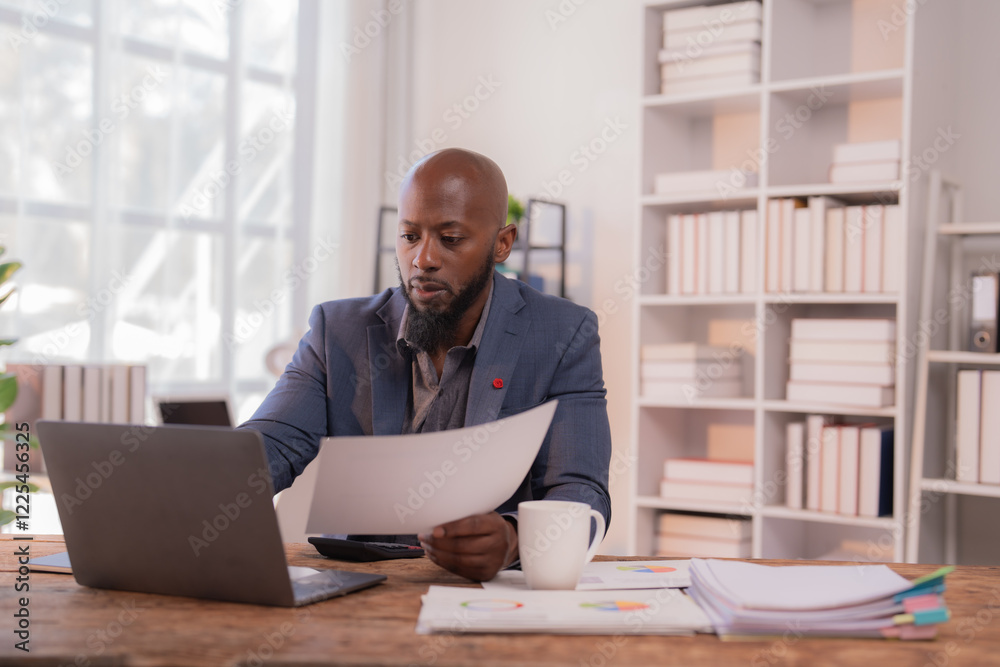 Businessman analyzing paperwork and using laptop, showcasing productivity and focus in a contemporary workspace with charts and graphs on the desk Businessman analyzing paperwork and using laptop, showcasing productivity and focus in a contemporary workspace with charts and graphs on the desk